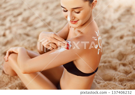 Suntan Lotion. Sexy Young Woman Applying Sunscreen Solar Cream on the beach.Sun Protection. Sun Cream. Skin and Body Care.Girl Holding Moisturizing Sunblock. Sun shape on the shoulder. Skin Protection 137406232