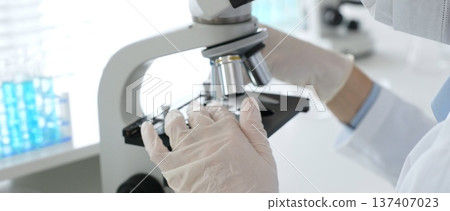 Gloved hands adjusting a microscope in a bright laboratory, emphasizing precision and focus in scientific research. Medicine, healthcare and science concept Gloved hands adjusting a microscope in a bright laboratory, emphasizing precision and focus in scientific research. Medicine, healthcare and science concept 137407023