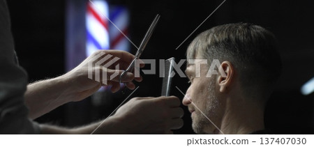 Professional barber carefully cutting hair of male customer with scissors, providing stylish and precise haircut in modern barbershop background 137407030