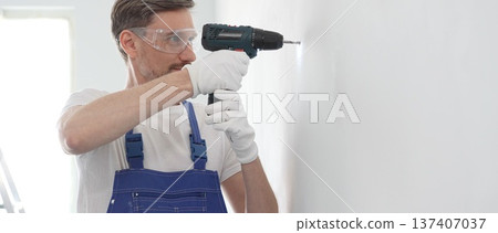 Middle aged man wearing blue overall and protective glasses, is drilling hole in white wall, wearing protective gear during home improvement project with cordless power tool Middle aged man wearing blue overall and protective glasses, is drilling hole in white wall, wearing protective gear during home improvement project with cordless power tool 137407037