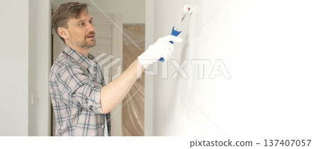 Professional male painter wearing checkered shirt, is rolling white paint on apartment wall, transforming interior space with steady strokes during comprehensive home renovation 137407057