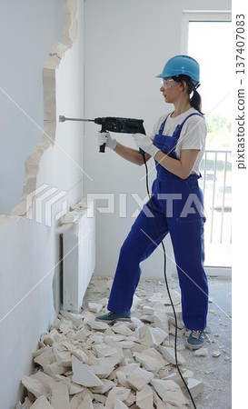 Female construction worker demolishing interior wall with rotary hammer, wearing safety glasses and hardhat, creating substantial concrete debris pile during demolition process 137407083