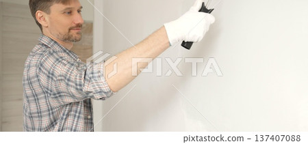 Man professional painter wearing checkered shirt, is rolling white paint on apartment wall with paint roller, transforming interior space with steady strokes during comprehensive home renovation Man professional painter wearing checkered shirt, is rolling white paint on apartment wall with paint roller, transforming interior space with steady strokes during comprehensive home renovation 137407088