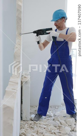 Construction worker demolishing concrete wall with rotary hammer, wearing protective safety gear, generating dust and debris during home renovation work 137407090