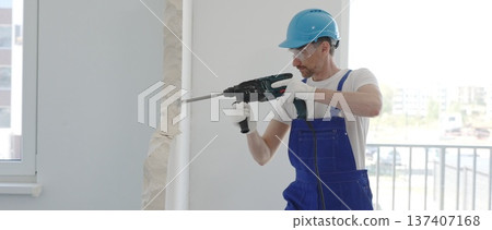 Male construction worker demolishing concrete wall with rotary hammer, wearing work overalls and blue hardhat is generating dust and debris during home renovation work, portrait horizontal view 137407168