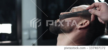 Skilled barber carefully combing thick beard of male client, providing professional grooming service in traditional barbershop setting with precision and attention to detail 137407172