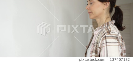 Middle aged woman wearing gloves is skillfully applying paint to a white wall with a paint roller in an apartment undergoing renovation, transforming the space with her creative touch Middle aged woman wearing gloves is skillfully applying paint to a white wall with a paint roller in an apartment undergoing renovation, transforming the space with her creative touch 137407182