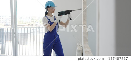 Female construction worker demolishing interior wall with rotary hammer, wearing safety glasses and hardhat, creating substantial concrete debris pile during demolition process Female construction worker demolishing interior wall with rotary hammer, wearing safety glasses and hardhat, creating substantial concrete debris pile during demolition process 137407217