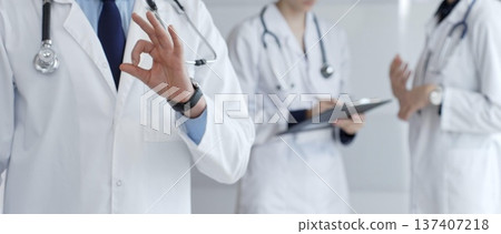 Professional doctor making an ok sign gesture, standing confidently while a medical team records patient data and discusses health matters in the background of a modern hospital Professional doctor making an ok sign gesture, standing confidently while a medical team records patient data and discusses health matters in the background of a modern hospital 137407218