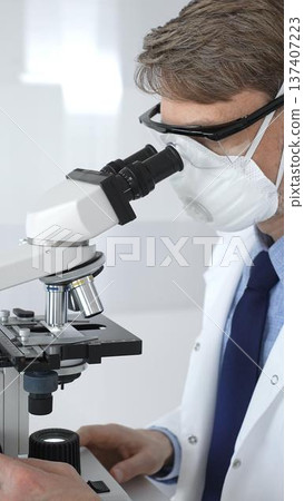A male scientist in a protective mask looks into a microscope in a brightly lit laboratory, conducting research to develop new drugs and treatments. Medicine and science concept 137407223