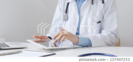 Female doctor wearing white medical coat and stethoscope using digital tablet with laptop and medical records on desk in medical office, showcasing modern healthcare technology. Medicine and helthcare 137407253