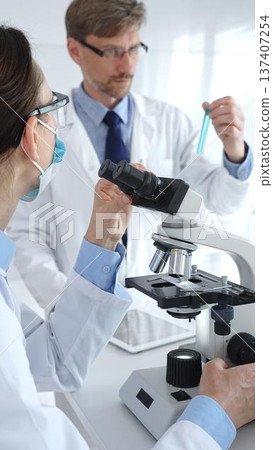 Two scientists in lab coats and safety gear conducting research, one observing through a microscope and the other holding a test tube with turquoise liquid. Medicine and science concept Two scientists in lab coats and safety gear conducting research, one observing through a microscope and the other holding a test tube with turquoise liquid. Medicine and science concept 137407254