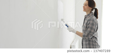 Middle aged happy woman wearing checked shirt and white gloves is painting white wall with paint roller, experiencing home renovation and transforming living space Middle aged happy woman wearing checked shirt and white gloves is painting white wall with paint roller, experiencing home renovation and transforming living space 137407289