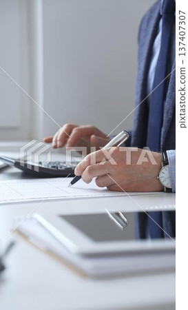 Businessman using calculator and taking notes recording financial data in office environment. Audit and taxes in business Businessman using calculator and taking notes recording financial data in office environment. Audit and taxes in business 137407307