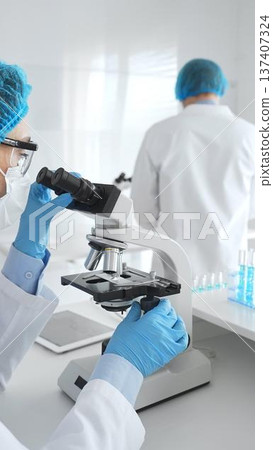Two scientists in lab coats, face masks, and protective glasses conducting research with a microscope in a laboratory. Medicine and science concept Two scientists in lab coats, face masks, and protective glasses conducting research with a microscope in a laboratory. Medicine and science concept 137407324