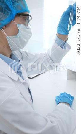 Medical science researcher wearing blue protective gear of cap, gloves and googles, is examining liquid in test tube in laboratory workspace. Medicine and health care concept 137407342
