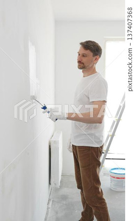 Middle aged man wearing white gloves and t-shirt, is painting white wall with paint roller, experiencing home renovation and transforming living space 137407368