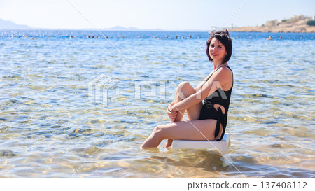 Elegant woman in a black swimsuit sitting on a plastic chair, enjoying the serene sea view, capturing the essence of summer relaxation and beach vibes 137408112