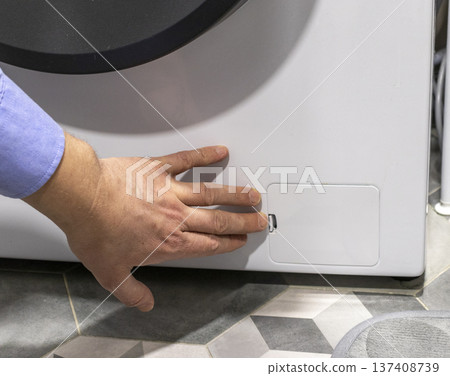Man opening the drain pump filter access panel of the front loading washing machine 137408739