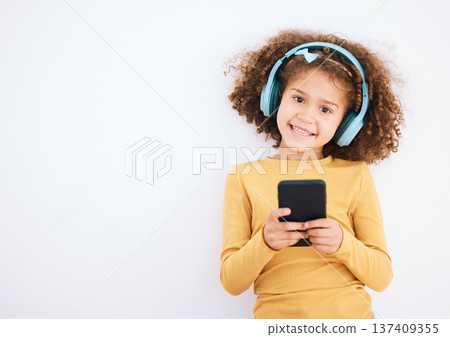 Girl child, portrait and smartphone, headphones and music with smile isolated on white background. Technology, audio streaming and social media, mockup space and kid, mobile app and radio in studio 137409355