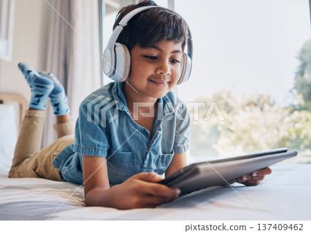 Boy, tablet and headphones on bed, reading of smile for streaming subscription, movie or relax with video. Male child, digital touchscreen and thinking in bedroom for audio, show or elearning in home Boy, tablet and headphones on bed, reading of smile for streaming subscription, movie or relax with video. Male child, digital touchscreen and thinking in bedroom for audio, show or elearning in home 137409462