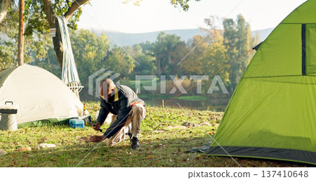 Camping, tent and black man setup in woods for shelter on outdoor holiday, vacation and adventure, Campsite, traveling and male person with hammer for pins for gear in nature, forest and countryside Camping, tent and black man setup in woods for shelter on outdoor holiday, vacation and adventure, Campsite, traveling and male person with hammer for pins for gear in nature, forest and countryside 137410648