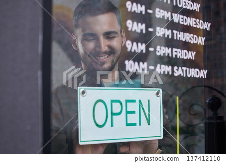 Shop, window and happy businessman open with sign for working time in retail, store or service in small business. Opening, restaurant or startup owner in Portugal with a welcome, poster and smile Shop, window and happy businessman open with sign for working time in retail, store or service in small business. Opening, restaurant or startup owner in Portugal with a welcome, poster and smile 137412110