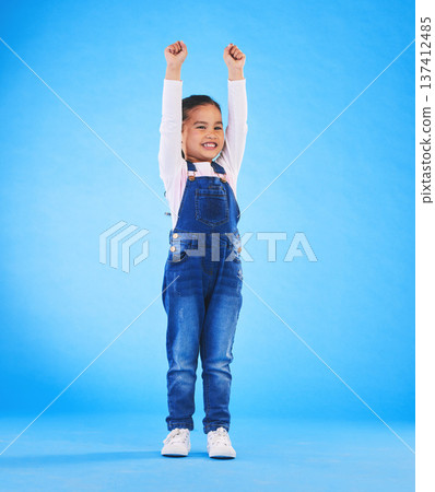 Celebration, girl child and champion, winner and achievement with happiness and excited isolated on blue background. Cheers, yes and winning with success, pride and happy kid with arms up in a studio Celebration, girl child and champion, winner and achievement with happiness and excited isolated on blue background. Cheers, yes and winning with success, pride and happy kid with arms up in a studio 137412485