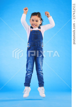 Jump, smile and portrait of a child in a studio with energy, achievement or good news with excitement. Happy, youth and full length of a girl kid model moving for winning by isolated blue background. 137412511