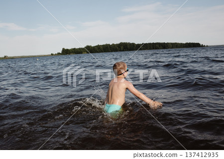 child happily excitedly enters water, swims, dives lake or pond on a hot, sunny summer day during vacation or school break. active healthy lifestyle. child wearing turquoise swimming trunks goggles. 137412935