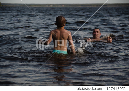 father and son swimming in a lake. man stands calmly in the water nearby. In the background is a green shore, calm waters, and a clear sky. The atmosphere evokes summer relaxation and family warmth. 137412951