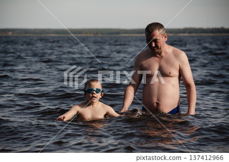 father and son swimming in a lake. man stands calmly in the water nearby. In the background is a green shore, calm waters, and a clear sky. The atmosphere evokes summer relaxation and family warmth. father and son swimming in a lake. man stands calmly in the water nearby. In the background is a green shore, calm waters, and a clear sky. The atmosphere evokes summer relaxation and family warmth. 137412966