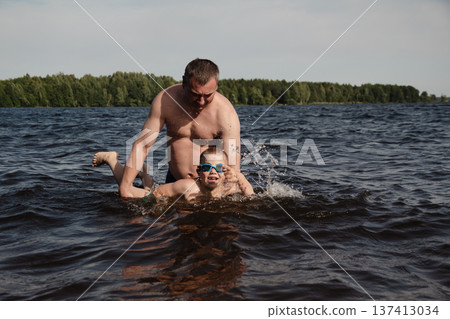 father and son swimming in a lake. man stands calmly in the water nearby. In the background is a green shore, calm waters, and a clear sky. The atmosphere evokes summer relaxation and family warmth. father and son swimming in a lake. man stands calmly in the water nearby. In the background is a green shore, calm waters, and a clear sky. The atmosphere evokes summer relaxation and family warmth. 137413034