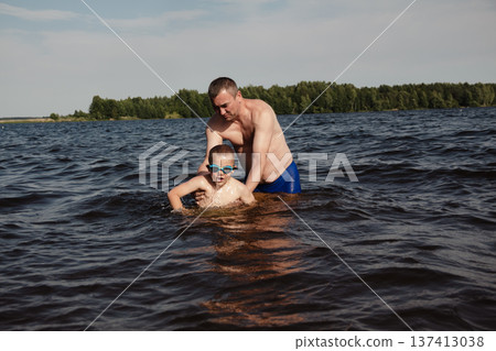 father and son swimming in a lake. man stands calmly in the water nearby. In the background is a green shore, calm waters, and a clear sky. The atmosphere evokes summer relaxation and family warmth. father and son swimming in a lake. man stands calmly in the water nearby. In the background is a green shore, calm waters, and a clear sky. The atmosphere evokes summer relaxation and family warmth. 137413038