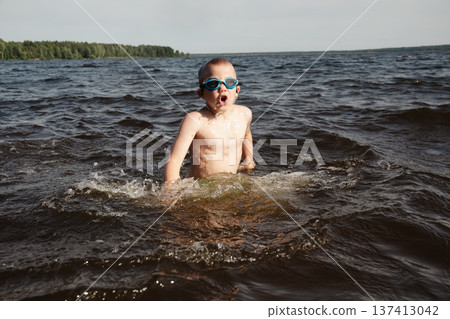 child happily excitedly enters water, swims, dives lake or pond on a hot, sunny summer day during vacation or school break. active healthy lifestyle. child wearing turquoise swimming trunks goggles. 137413042