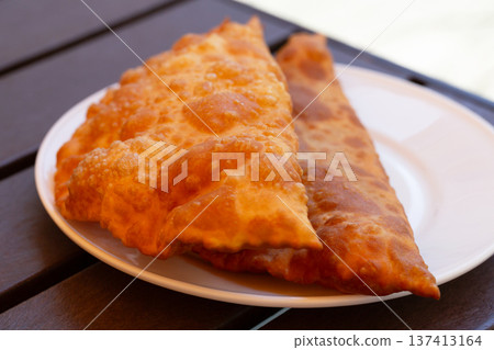Hearty lunch: Chebureks with meat on a white plate on a wooden table. Big chebureks for dinner. Meat pies. Close-up. Hearty lunch: Chebureks with meat on a white plate on a wooden table. Big chebureks for dinner. Meat pies. Close-up. 137413164