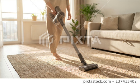 Person vacuums carpet in bright living room with sunlight coming through windows. Indoor setting features plants and cozy couch. Concept of home cleaning, interior design, housekeeping. Person vacuums carpet in bright living room with sunlight coming through windows. Indoor setting features plants and cozy couch. Concept of home cleaning, interior design, housekeeping. 137413402