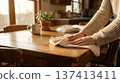Person wipes down wooden table in kitchen with sunlight streaming through windows. Scene includes houseplant and mug beside open book. Concept of home cleaning, organization, interior design. 137413411