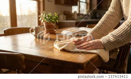 Person wipes down wooden table in kitchen with sunlight streaming through windows. Scene includes houseplant and mug beside open book. Concept of home cleaning, organization, interior design. Person wipes down wooden table in kitchen with sunlight streaming through windows. Scene includes houseplant and mug beside open book. Concept of home cleaning, organization, interior design. 137413411