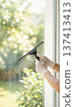 Person cleans window with squeegee in bright setting. Sunlight shines through, revealing green scenery outside. Concept of home cleaning, window care, residential services. 137413413