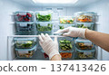 Hand wearing glove arranges clear containers filled with colorful vegetables and fruits in refrigerator. Clean kitchen setting, organized space. Concept of meal prep, health food, cooking. 137413426