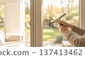 Person uses squeegee to clean glass window in living room with a view of greenery outside. Clear sunlight shines in through window. Concept of cleaning, home improvement, interior design. 137413462