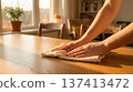 Person wipes wooden table with cloth in cozy room filled with sunlight. Bright setting with soft lighting and furniture in background. Concept of cleaning, home care, interior design. 137413472