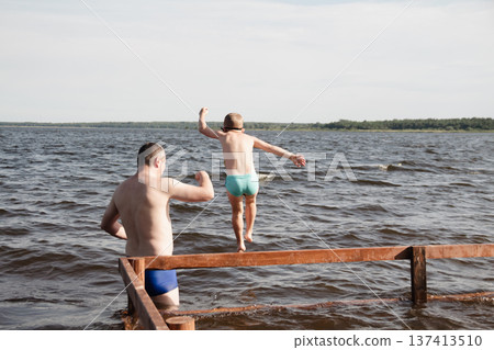 father and son swimming in a lake. man stands calmly in the water nearby. In the background is a green shore, calm waters, and a clear sky. The atmosphere evokes summer relaxation and family warmth. father and son swimming in a lake. man stands calmly in the water nearby. In the background is a green shore, calm waters, and a clear sky. The atmosphere evokes summer relaxation and family warmth. 137413510