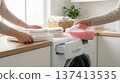Two people fold clean towels while one pours pink liquid detergent into washing machine. Setting in bright kitchen with natural light and plants. 137413535