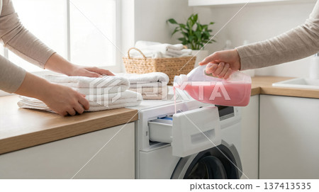 Two people fold clean towels while one pours pink liquid detergent into washing machine. Setting in bright kitchen with natural light and plants. Two people fold clean towels while one pours pink liquid detergent into washing machine. Setting in bright kitchen with natural light and plants. 137413535