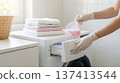 Person pours pink liquid detergent into drawer of washing machine. Laundry room setting with organized towels on top of machine. Concept of laundry services, home cleaning, household tasks. 137413544