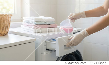 Person pours pink liquid detergent into drawer of washing machine. Laundry room setting with organized towels on top of machine. Concept of laundry services, home cleaning, household tasks. Person pours pink liquid detergent into drawer of washing machine. Laundry room setting with organized towels on top of machine. Concept of laundry services, home cleaning, household tasks. 137413544