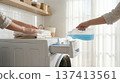 Person pours blue laundry detergent into washing machine while folding clean clothes on countertop. Clean and bright laundry room with white tiles. Concept of cleaning, home care, household services. 137413561