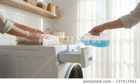 Person pours blue laundry detergent into washing machine while folding clean clothes on countertop. Clean and bright laundry room with white tiles. Concept of cleaning, home care, household services. Person pours blue laundry detergent into washing machine while folding clean clothes on countertop. Clean and bright laundry room with white tiles. Concept of cleaning, home care, household services. 137413561